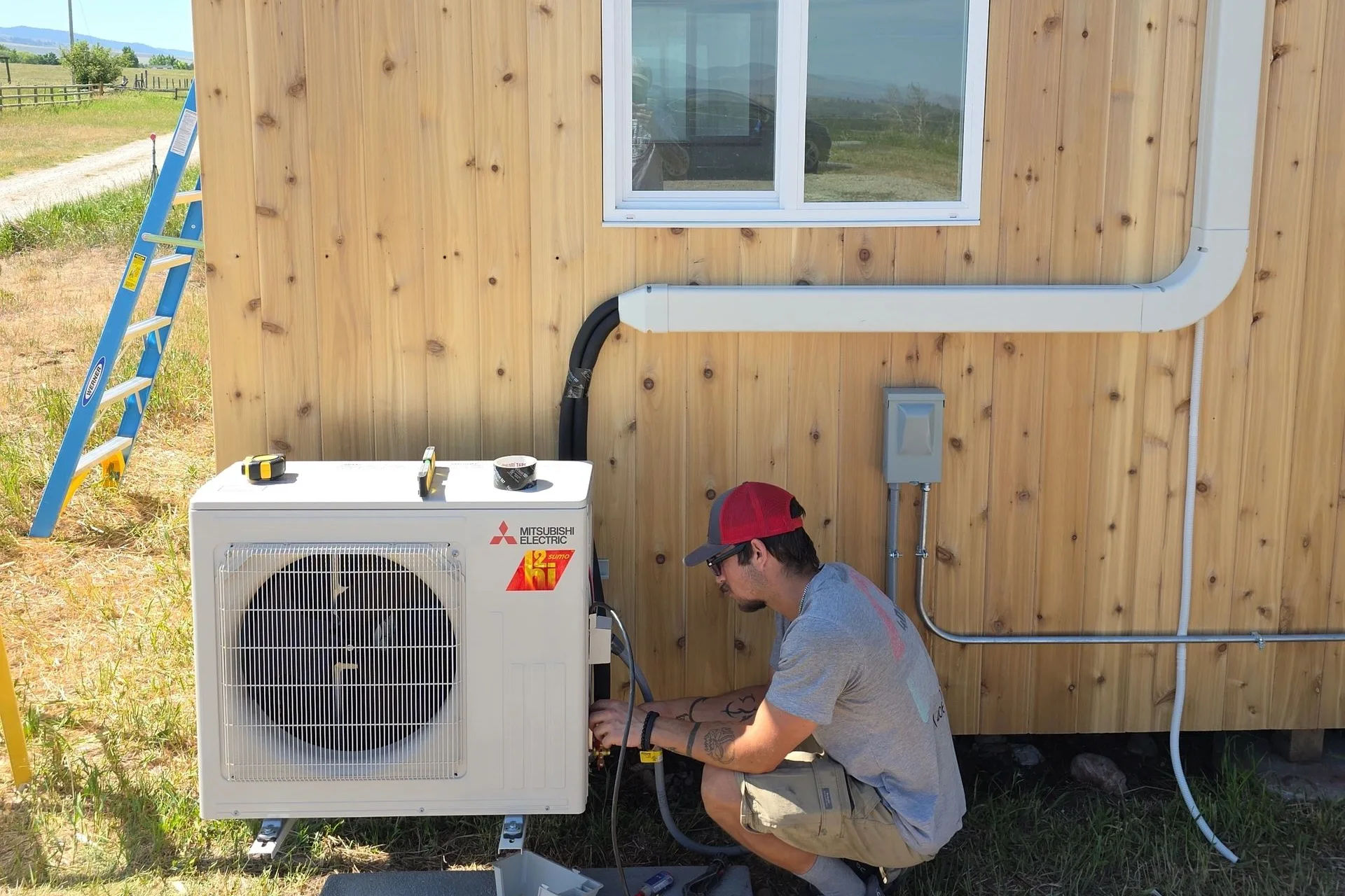 406 Heating and Cooling technician charging a Mitsubishi Electric mini-split outdoor unit at a cedar-sided home in the Missoula valley