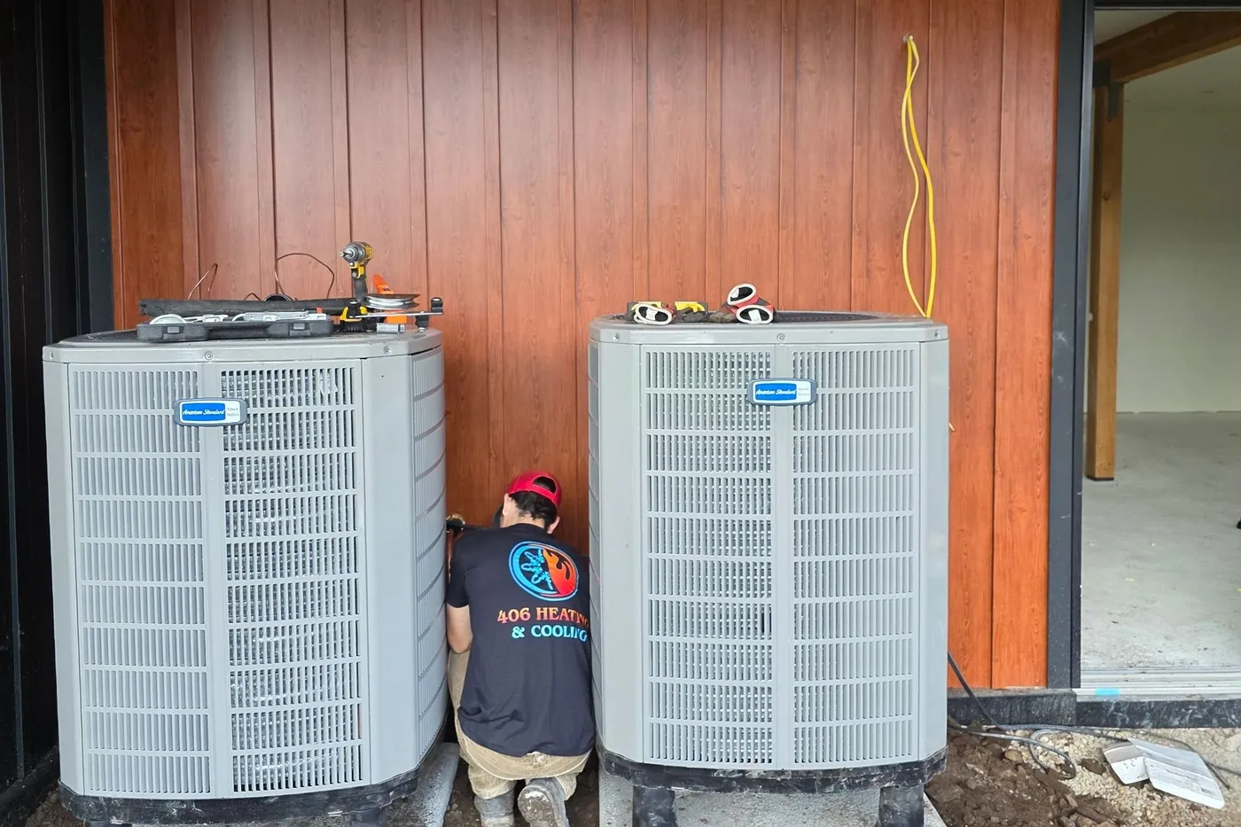 406 Heating and Cooling technician in a branded company shirt working between two American Standard condensers at a new construction home