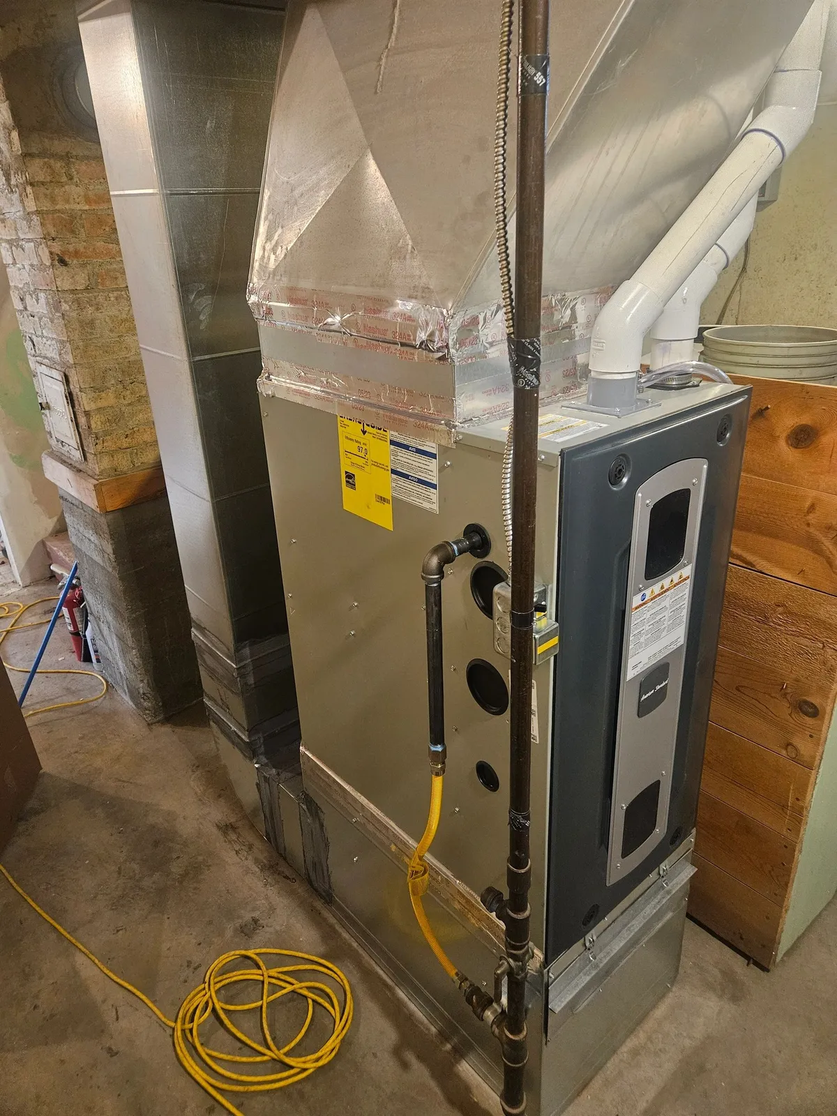 High-efficiency American Standard gas furnace in a basement with a newly fabricated sheet metal supply plenum and taped transition above it, PVC venting run to the wall and a yellow flexible gas connector at the base