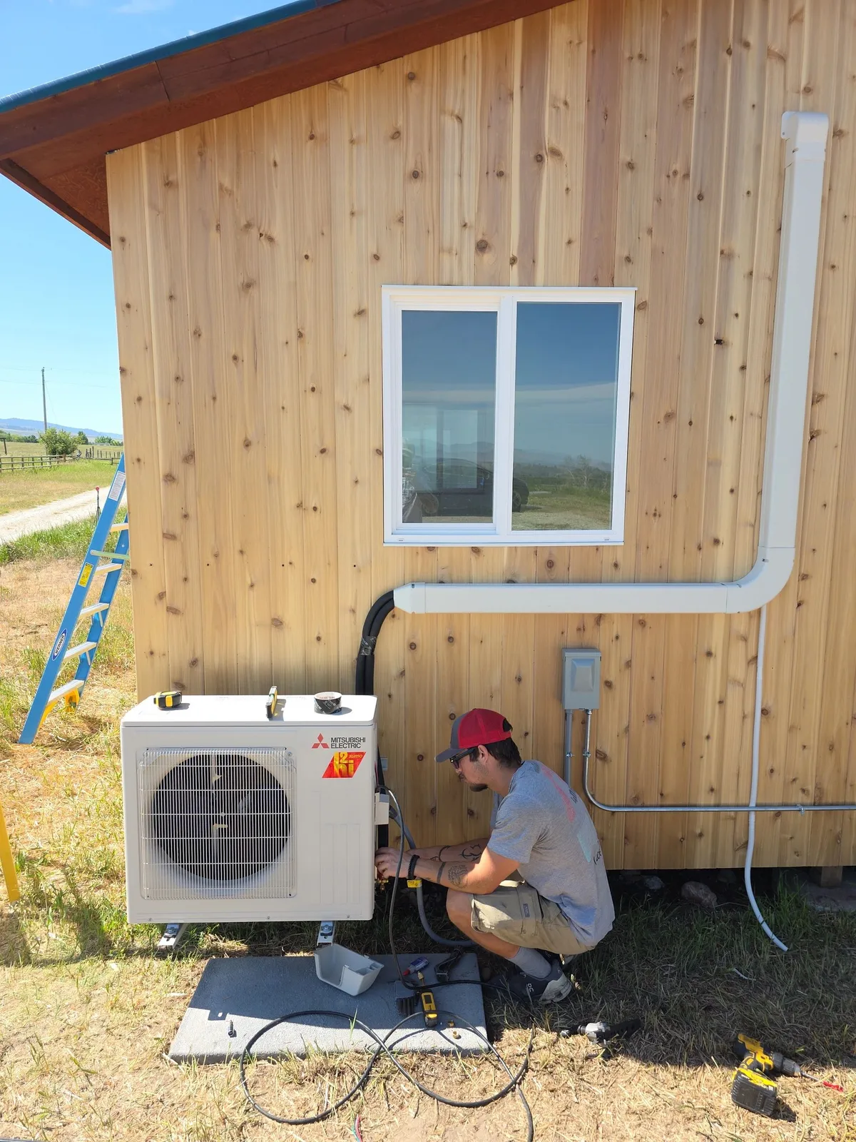 406 Heating and Cooling technician kneeling to charge a Mitsubishi Electric mini-split outdoor unit mounted on a pad against the cedar siding of a new Montana build, with gauges connected and a ladder leaning nearby