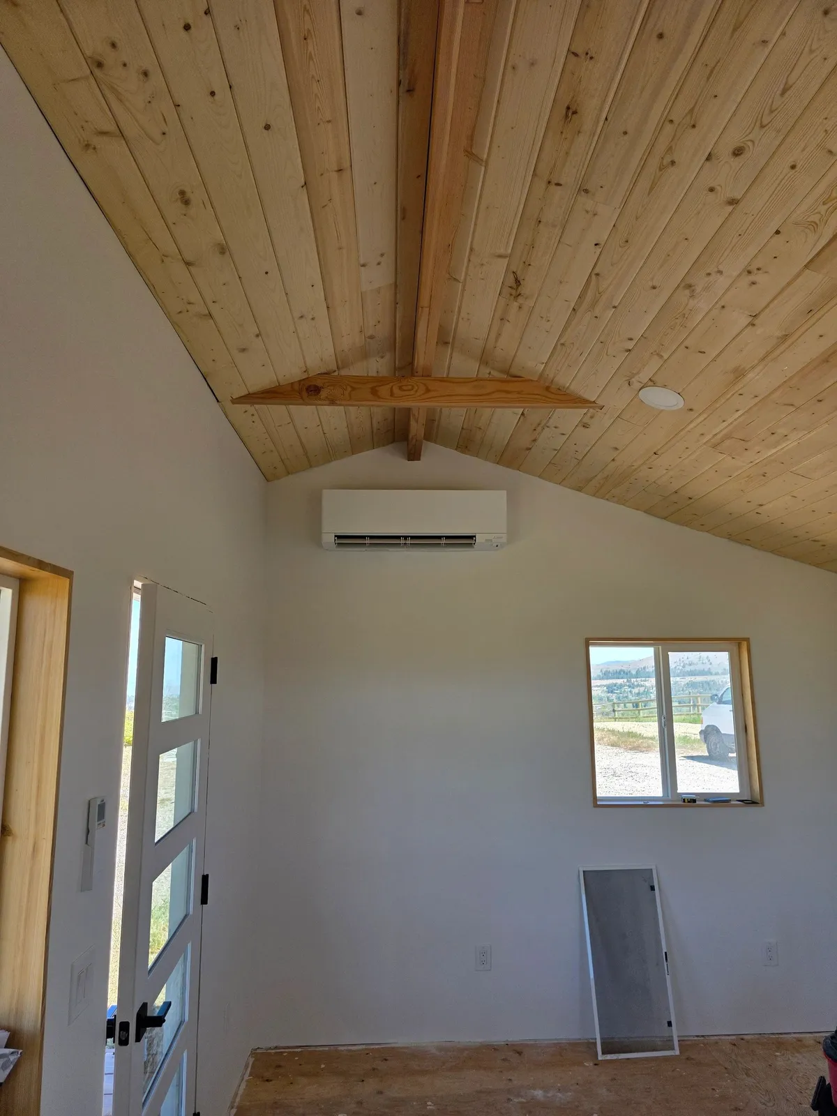 Mitsubishi Electric wall-mounted mini-split head installed high on a white wall beneath a vaulted cedar plank ceiling, with a window looking out to Montana hills