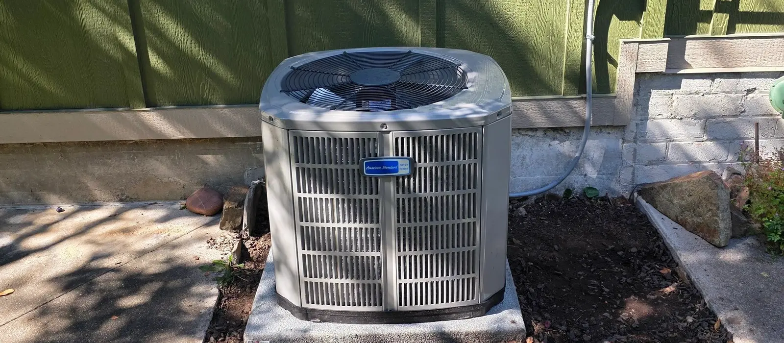 American Standard air conditioning condenser installed on a concrete pad beside a green-sided home with the electrical disconnect mounted on the wall above
