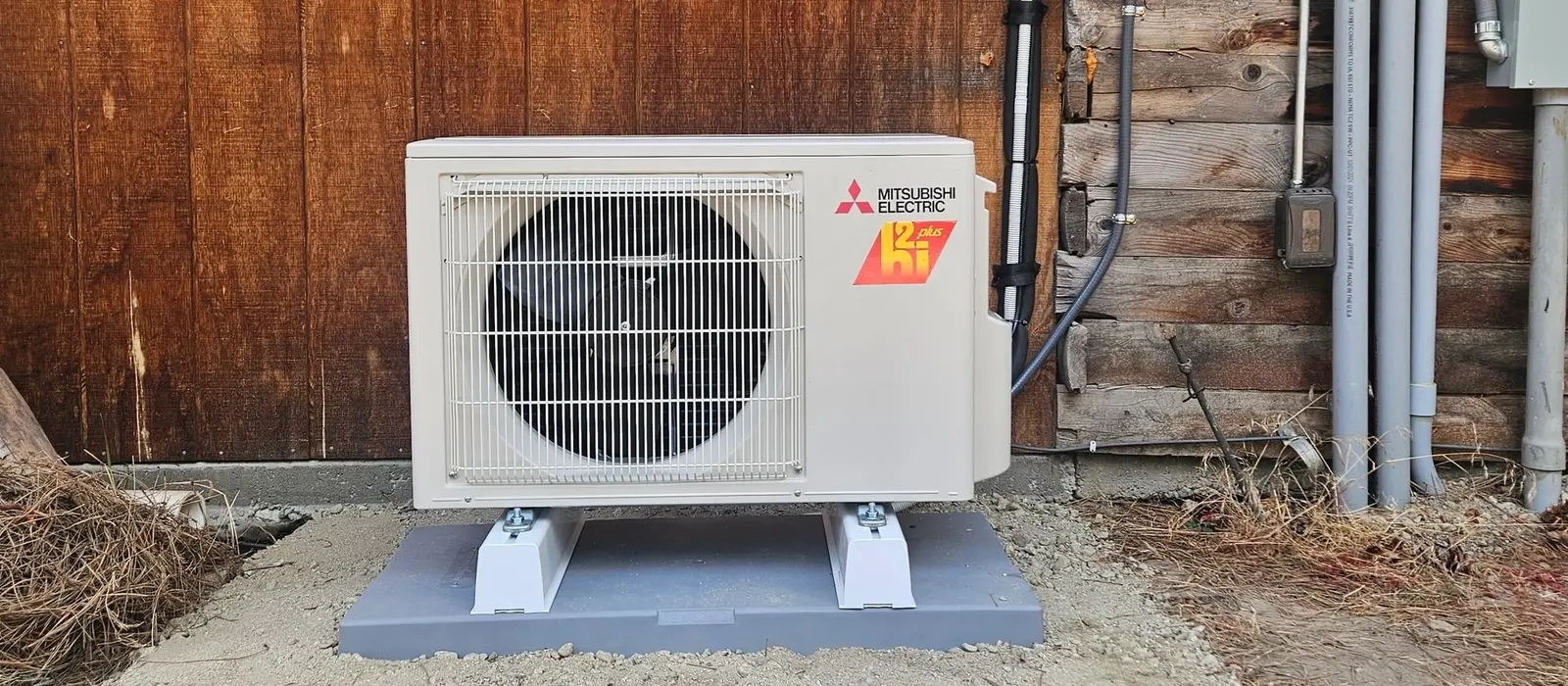 Mitsubishi Electric outdoor heat pump unit mounted on a grey pad against the log wall of a Montana home, with electrical disconnect and conduit run above