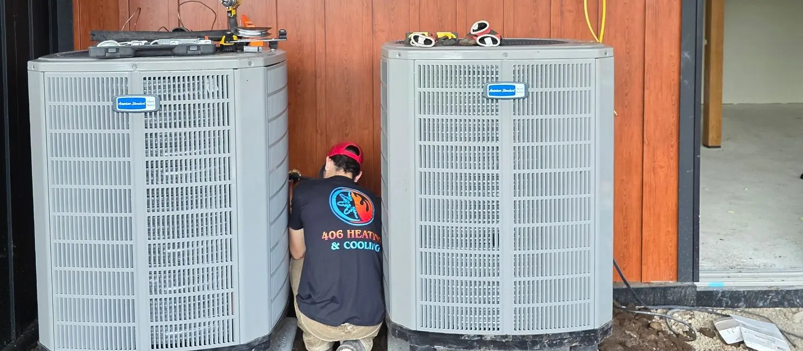 Two American Standard condensers set on concrete pads at a new construction home with a 406 Heating and Cooling technician in a branded company shirt working between them