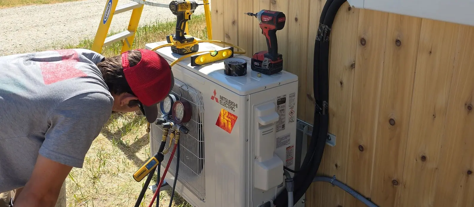 406 Heating and Cooling technician charging a Mitsubishi Electric mini-split outdoor unit with refrigerant gauges attached, tools resting on the cabinet and open Montana country behind