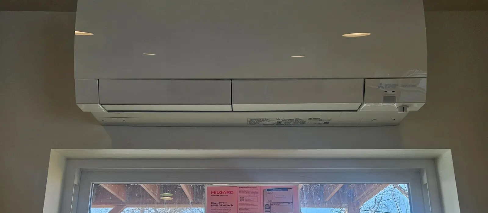 Mitsubishi Electric wall-mounted mini-split head installed above a window, with bare winter trees and snow on the roofline visible outside