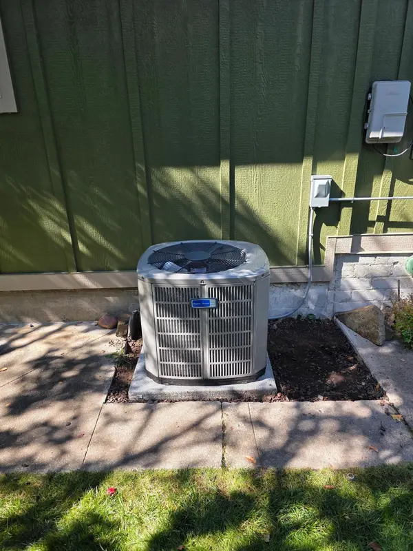 American Standard air conditioning condenser installed on a concrete pad beside a green-sided home, with the electrical disconnect and whip mounted on the wall above and a fresh mulch bed alongside