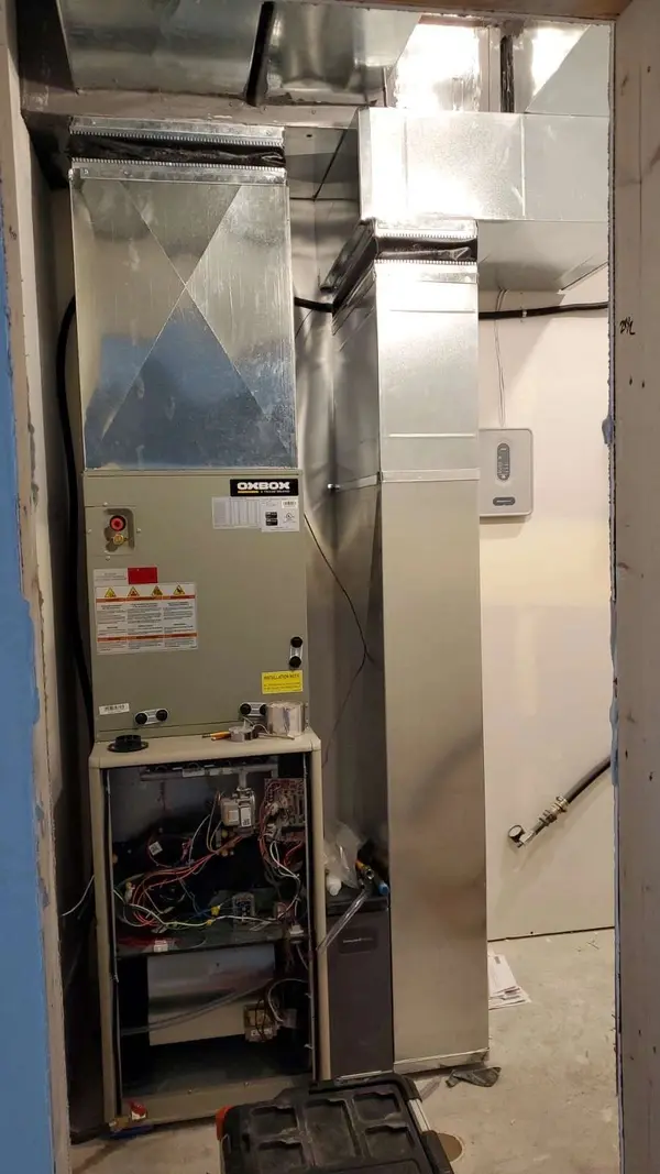Air handler and newly fabricated sheet metal supply trunk installed in the mechanical closet of a home under construction, with the return drop beside it and a thermostat wired to the unfinished wall