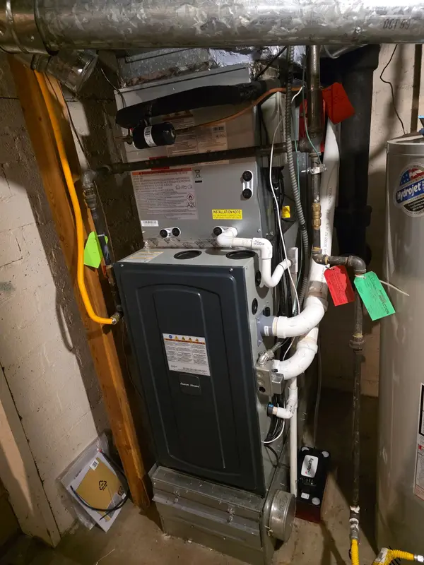 American Standard gas furnace with the evaporator coil cased above it in a basement, gas piping and condensate lines run, and a green approved-inspection tag hanging beside a red tag on the pipework
