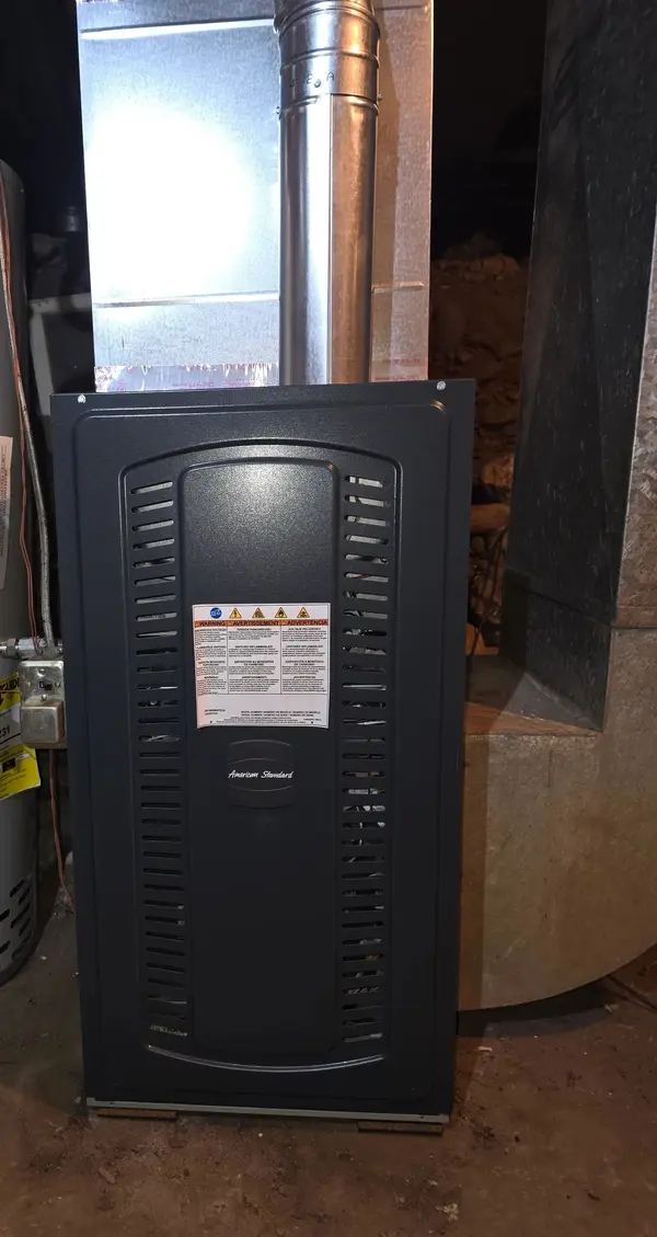 Newly installed American Standard gas furnace standing on a sheet metal base in a basement, with the flexible gas connector and shutoff made up at the side