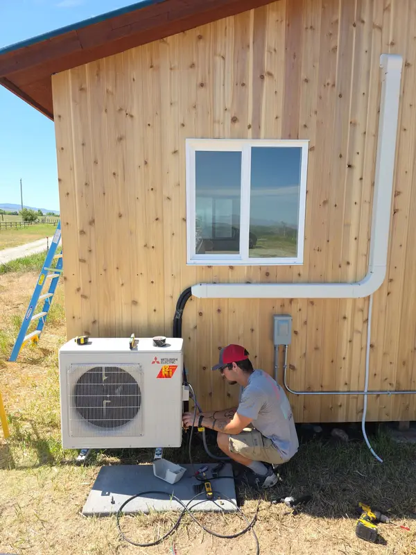 406 Heating and Cooling technician kneeling to charge a Mitsubishi Electric mini-split outdoor unit mounted on a pad against the cedar siding of a new Montana build, with gauges connected and a ladder leaning nearby