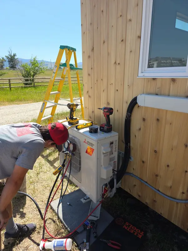 Technician bent over a Mitsubishi Electric mini-split outdoor unit with refrigerant gauges attached, tools and a level resting on the cabinet, open Montana pasture and hills behind