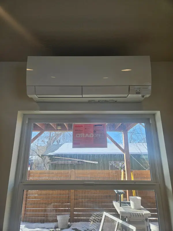 Mitsubishi Electric wall-mounted mini-split head installed above a window, with snow on the roofline and bare trees visible outside