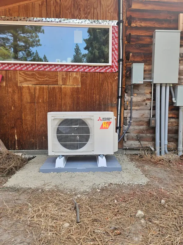 Mitsubishi Electric outdoor unit set on a grey composite pad against the log wall of a Montana cabin, with electrical disconnect, meter base and conduit runs on the wall alongside