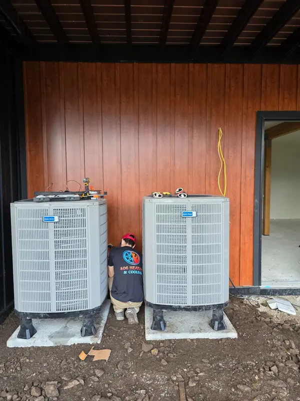 Two American Standard condensers set on concrete pads on equipment feet at a new construction home with wood panel siding, and a 406 Heating and Cooling technician in a branded company shirt working between them